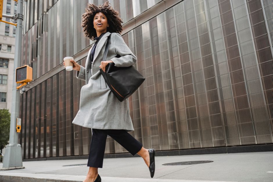 Low angle of confident African American female in trendy outfit with bag holding coffee while crossing road in downtown of city