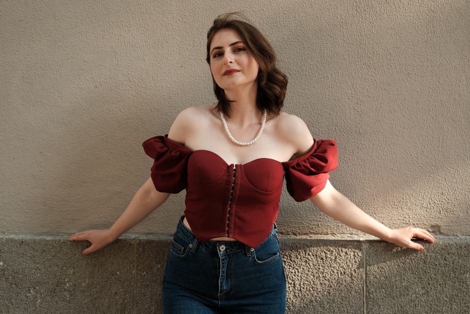 Stylish woman in off-shoulder red blouse poses confidently against a textured wall.