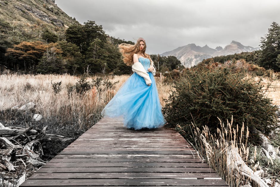 A woman in a flowing blue gown poses on a boardwalk surrounded by nature, exuding elegance.