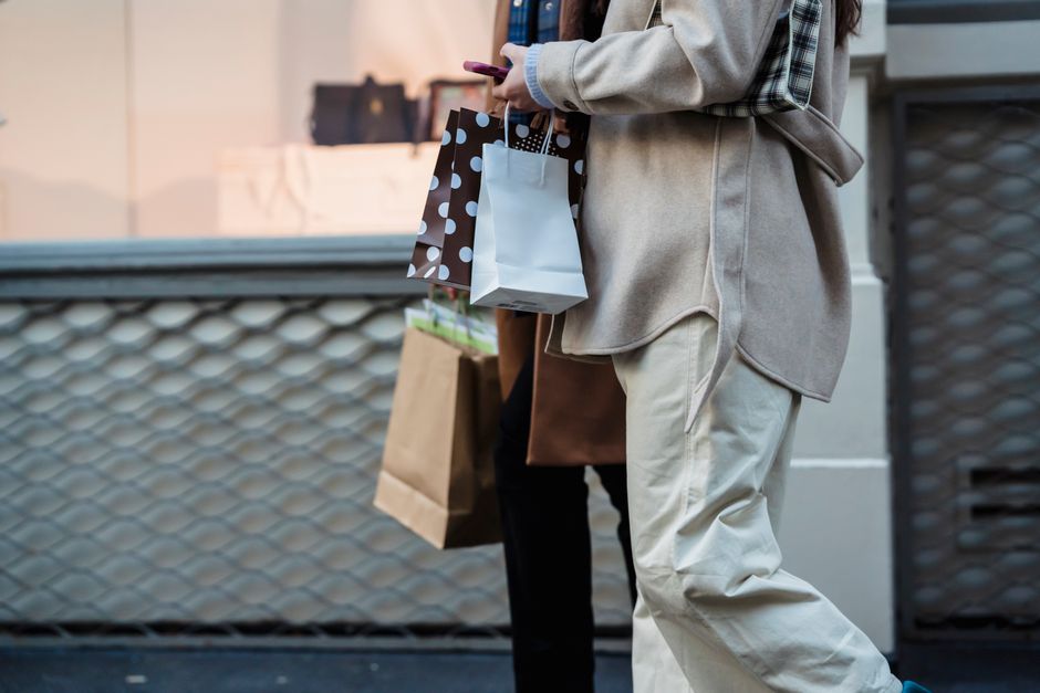 Side view of crop unrecognizable female shopaholics in trendy outfits with paper bags walking together on street after shopping and using smartphone