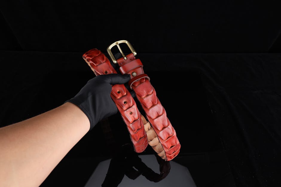 A red leather belt with a gold buckle is elegantly held by a gloved hand against a black background.