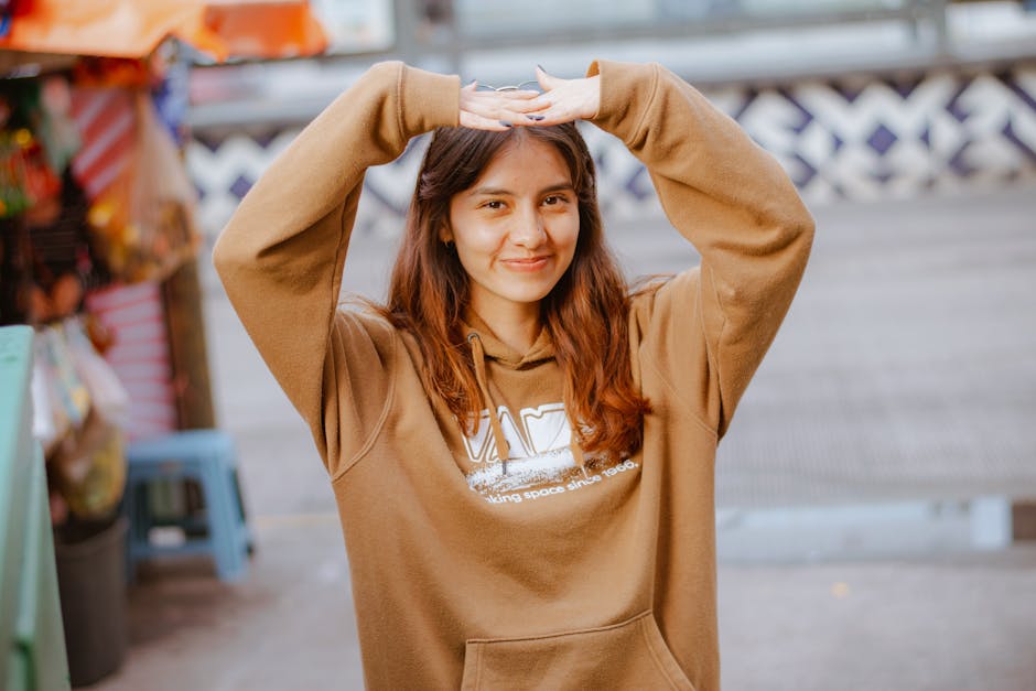 A young woman smiling and posing playfully outdoors in a casual brown hoodie. Warm and joyful moment.