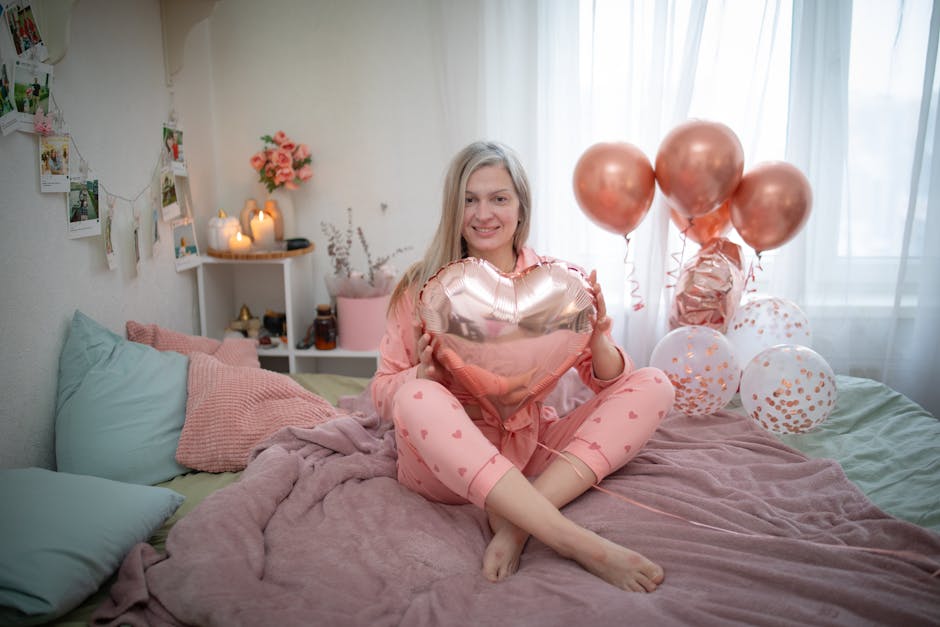 A blonde woman in pajamas sits on a bed holding a heart balloon, surrounded by a cozy bedroom atmosphere.