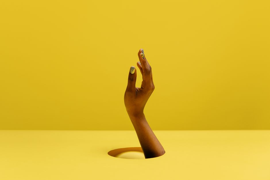 Close-up of a hand with manicure emerging from a yellow background, signifying creativity.