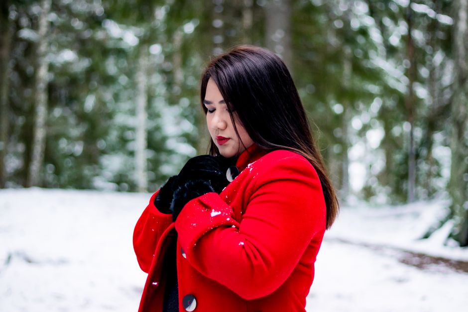 A woman in a vibrant red coat stands thoughtfully in a snowy forest in Finland.