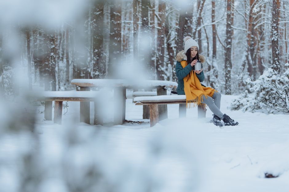 Asian woman in winter attire sipping a drink in a snowy forest. Serene and cosy atmosphere.