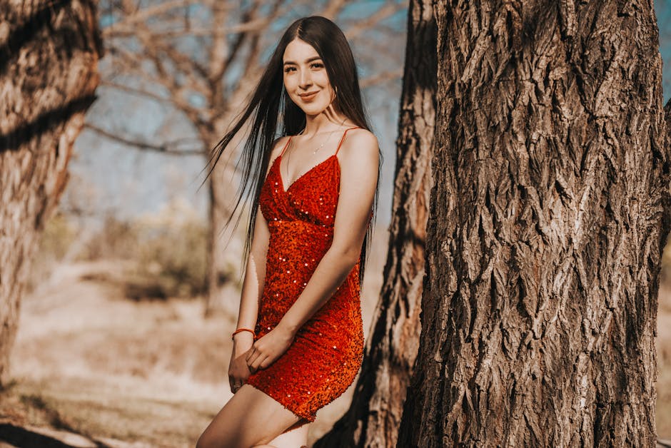 A woman in a sparkling red dress leans against a tree in a rustic outdoor setting.