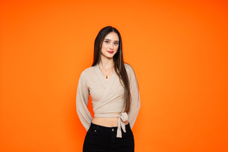 A young woman with long hair smiling confidently in casual attire against a vibrant orange background.