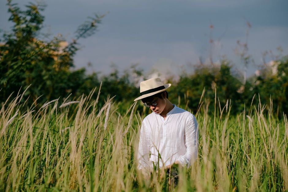 A fashionable man in a white shirt and hat stands in a lush green field under a clear blue sky.