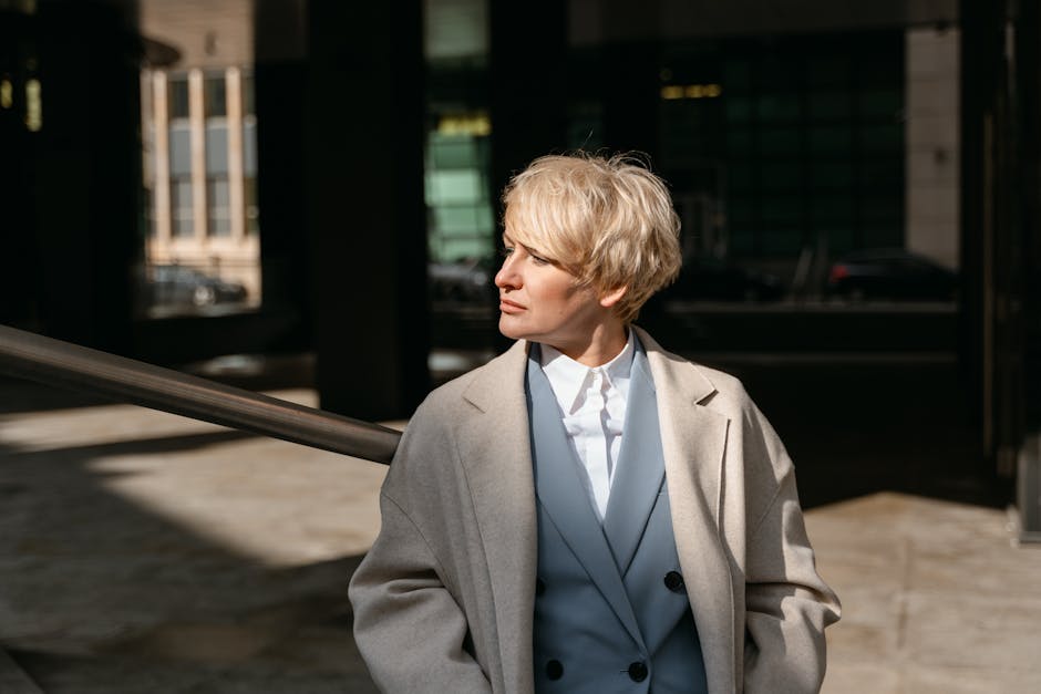Professional blonde woman in a coat, facing away in a sunlit urban environment.