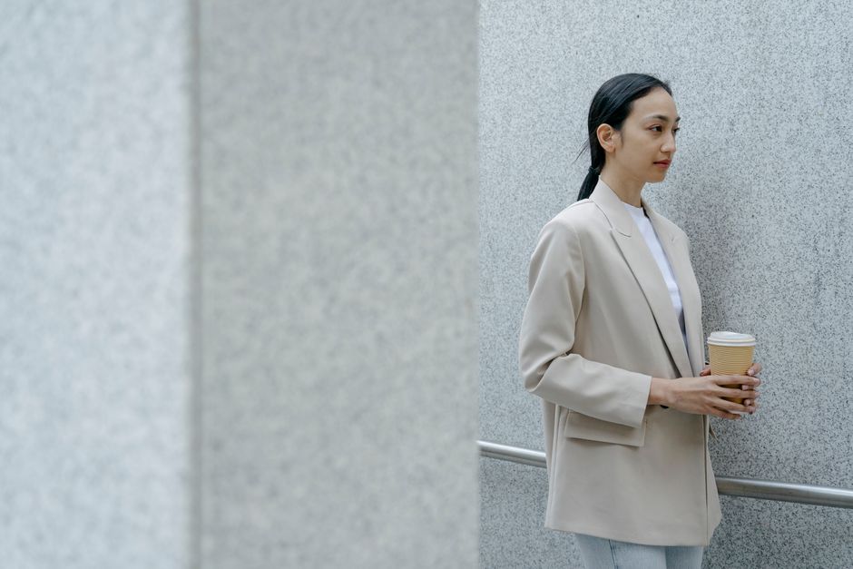 Emotionless Asian female in formal outfit with cup of hot drink standing near railing and looking away