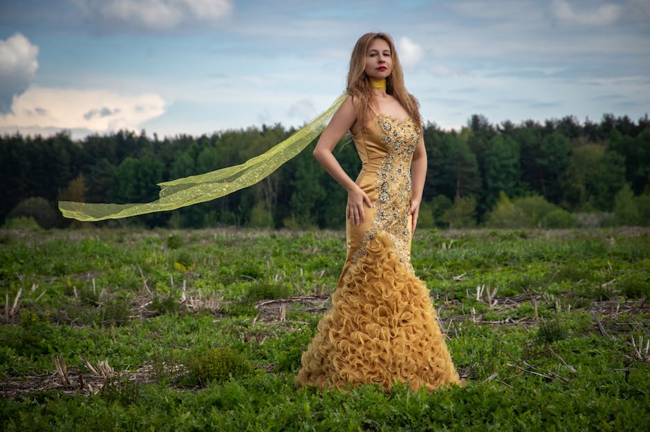 A woman in a golden evening gown stands confidently in an open field under a cloudy sky.