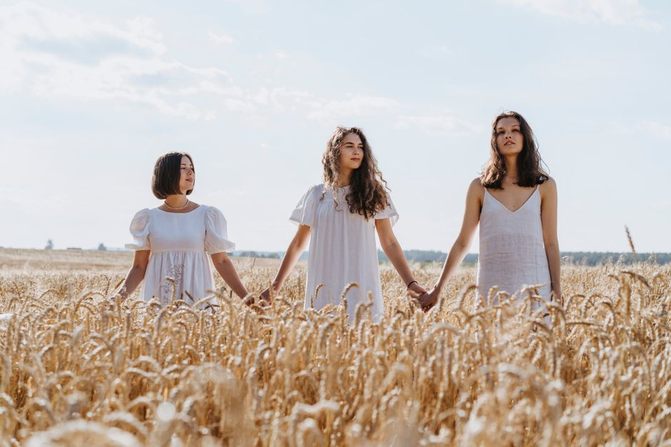 Three women in white dresses holding hands in a sunlit wheat field.