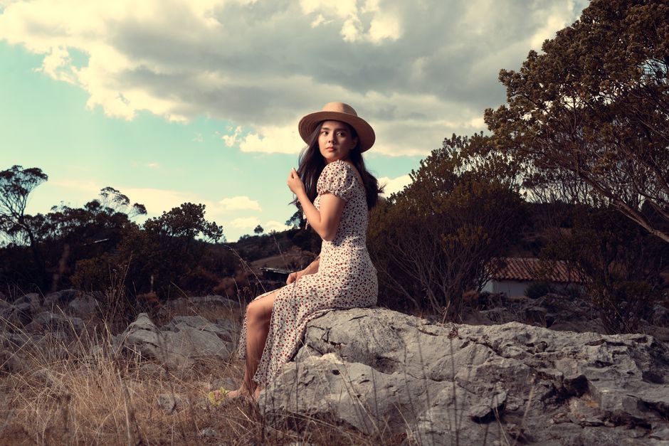 Woman in floral dress and hat sitting on rocky terrain under cloudy skies.