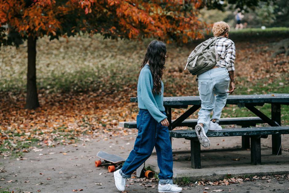 Full body of unrecognizable female with dark hair and African American partner with backpack jumping on bench resting in autumn nature