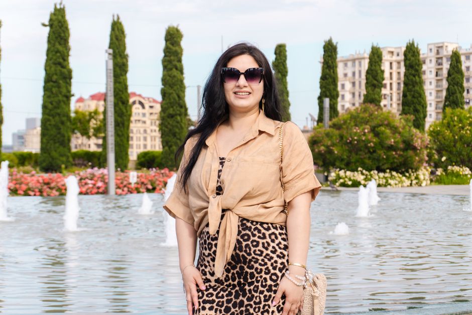 Stylish woman in leopard print skirt posing by a city fountain surrounded by tall trees and buildings.