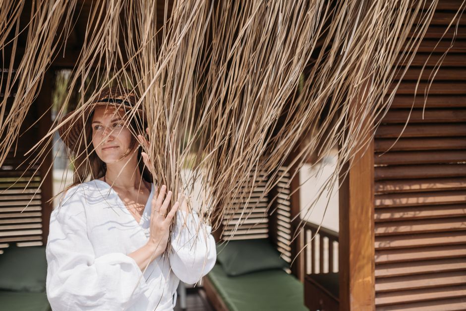 A woman enjoys a peaceful moment under a thatched beach hut in a sunny tropical setting.