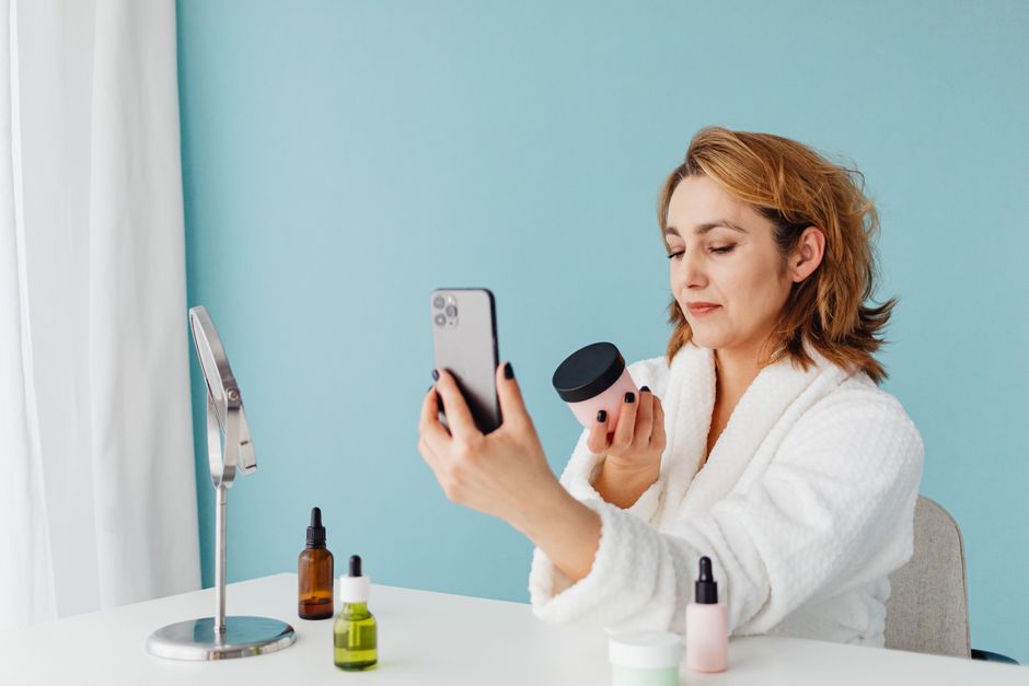 Woman in a white robe taking a selfie with skincare products, set against a light blue background.