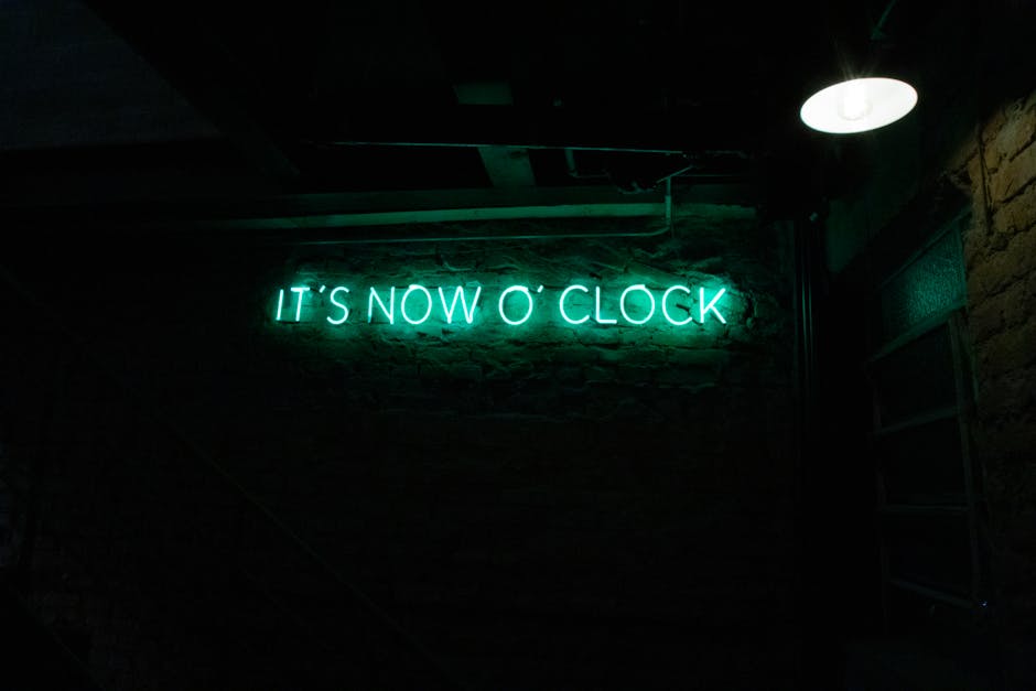 Green neon sign in a dimly lit room showcasing 'It's Now O' Clock'.