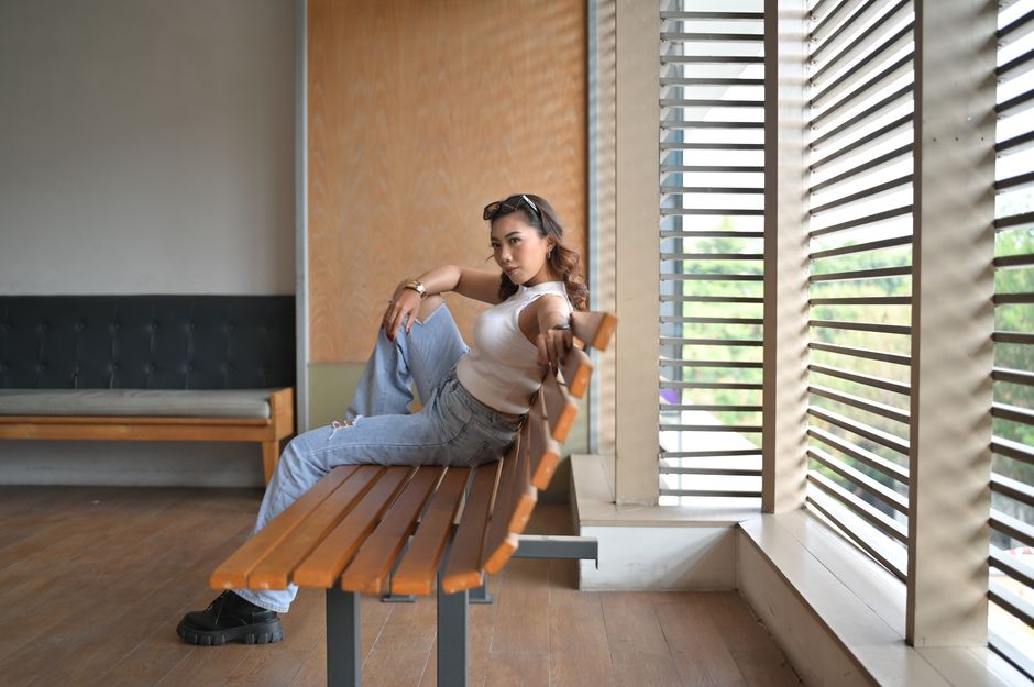 Stylish woman lounging on a bench indoors, Jakarta. Casual jeans and top fashion, bright ambiance.