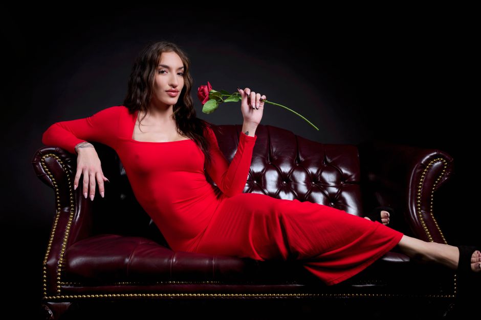 A woman in a red dress lies elegantly on a leather sofa holding a rose.
