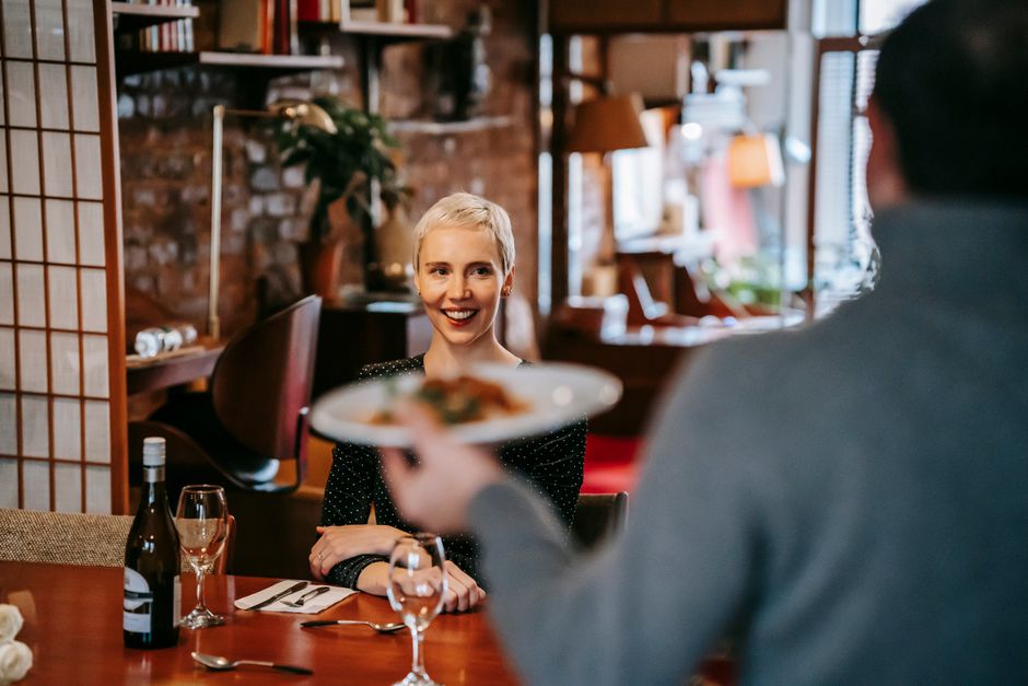 Positive couple having date in bright restaurant while man serving plate with food to woman sitting in chair at table with wine bottle and glasses near cutlery with napkin