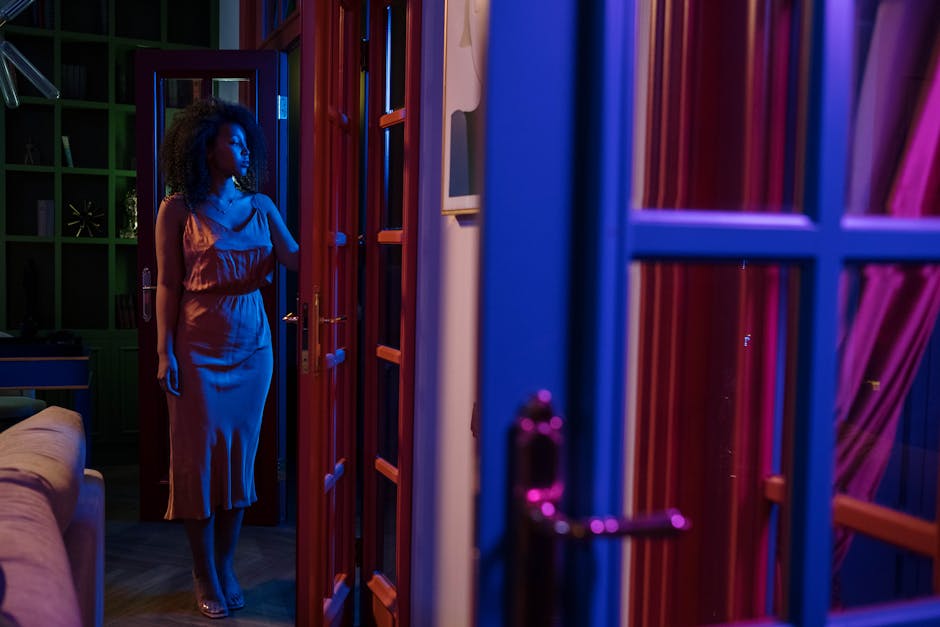 A woman in a satin dress stands by an open doorway at night lit in colorful tones.