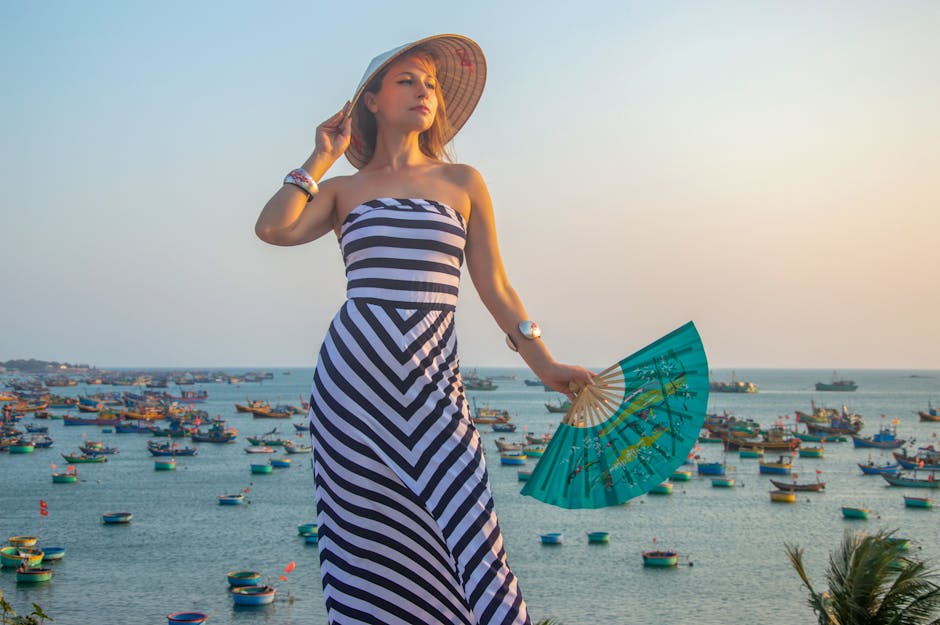Stylish woman in striped dress with fan stands by a colorful sea full of boats at sunset.